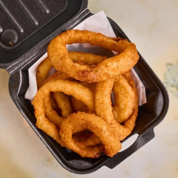Golden-brown, crispy onion rings are piled high in a black takeout container from GutBusters/VeggieBusters.