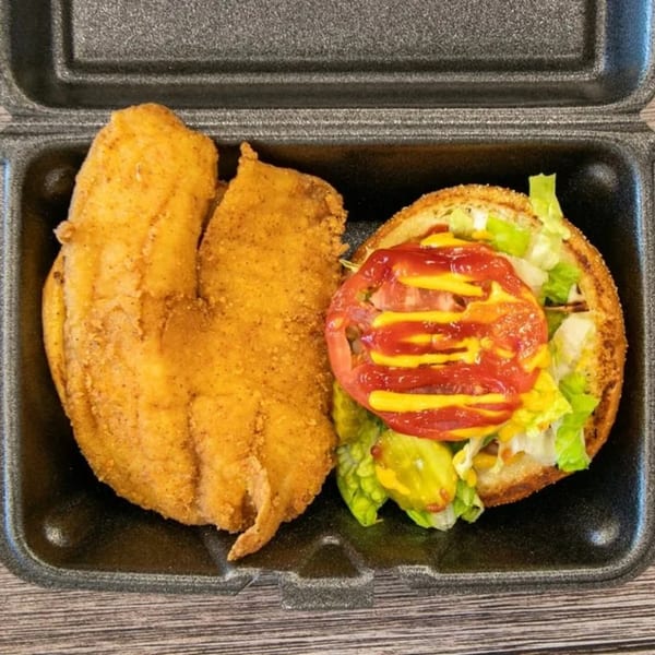 A GutBusters fish sandwich with lettuce, tomato, pickles, ketchup, and mustard, next to a fried fish fillet.