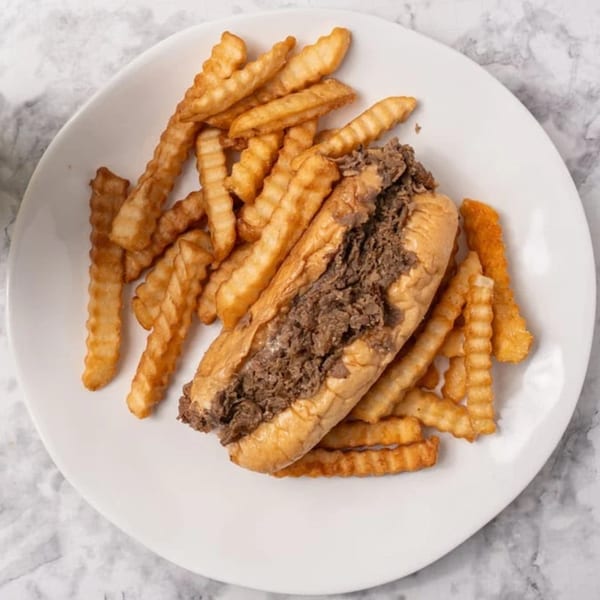 A GutBusters cheesesteak sandwich with crinkle-cut fries on a white plate.